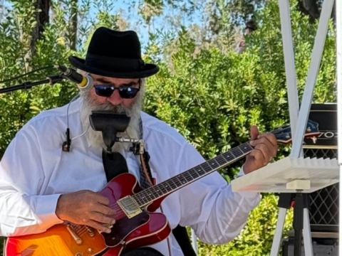 Bearded musician with a fedora and sunglasses plays a red hollow-body electric guitar and harmonica outdoors.
