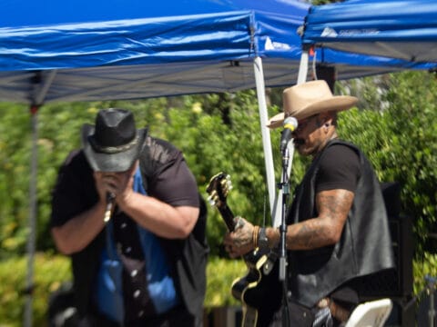Two musicians in cowboy hats perform under a blue canopy, one playing an electric guitar and the other playing harmonica.