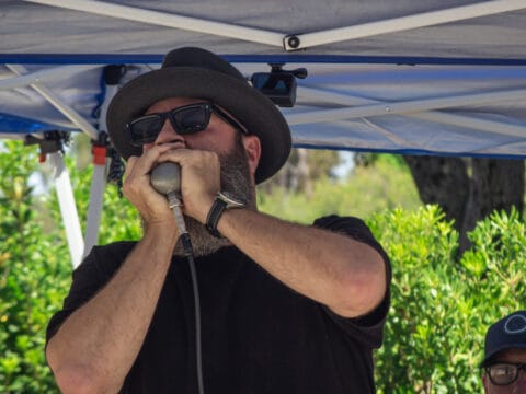 Bearded man in a fedora and sunglasses playing a harmonica into a microphone under a blue canopy.