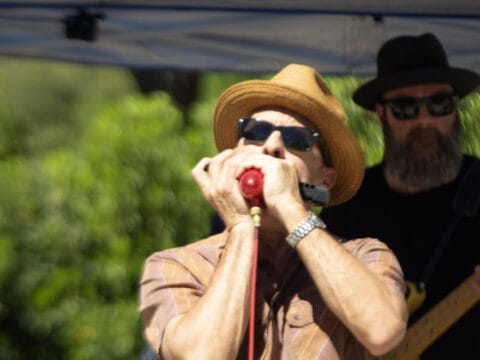 Musician wearing a tan fedora and sunglasses plays a harmonica into a red microphone outdoors.