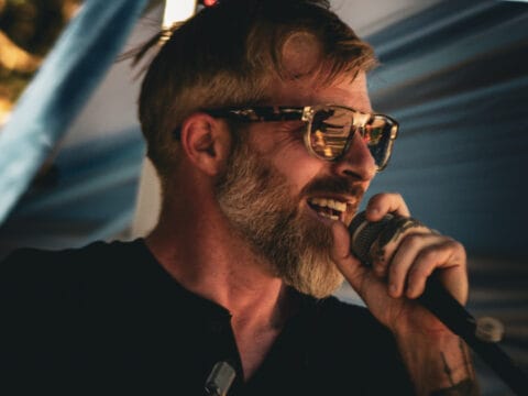 Bearded man with sunglasses singing into a microphone on stage under a canopy.