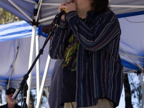 Musician wearing a brown hat and sunglasses plays the harmonica into a microphone under a blue tent canopy.
