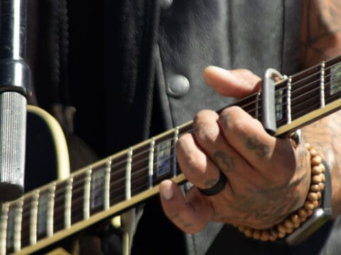 Tattooed hand with a black ring and wooden bead bracelet playing an acoustic guitar with a capo on the neck.