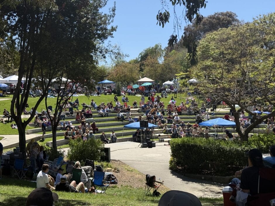 Crowd of people sitting on tiered concrete steps and grass in a sunny park at an outdoor event under blue pop-up tents.