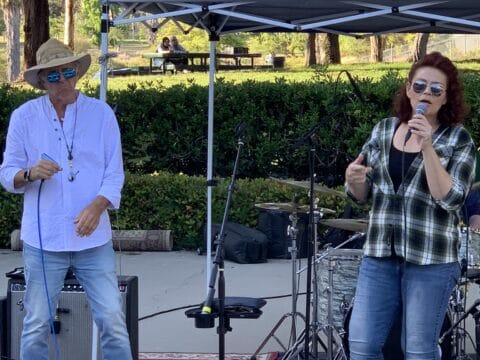 Man in a fedora and woman in a plaid shirt performing music together under a blue canopy in a park.