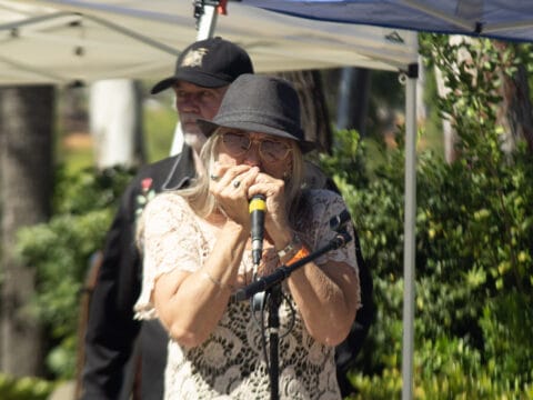 Woman wearing a fedora and sunglasses plays a harmonica into a microphone while a man in a baseball cap stands behind her.