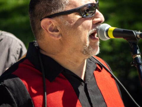 Middle-aged man with sunglasses singing into a microphone while wearing a black and red collared shirt outdoors.
