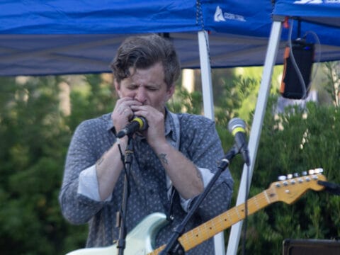 Musician playing harmonica into a microphone while holding an electric guitar under a blue canopy.