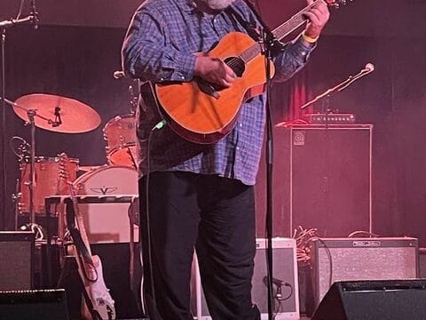 Bearded musician in a plaid shirt plays an acoustic guitar while singing into a microphone on a stage with concert lights
